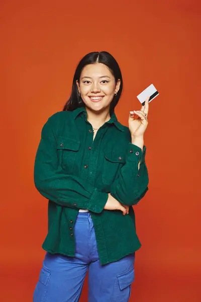 Heureux jeune asiatique femme en vert veste tenant carte de crédit sur fond orange, consumérisme — Photo de stock