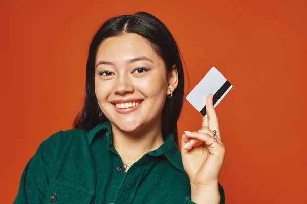 Feliz joven asiático mujer en verde chaqueta celebración de tarjeta de crédito en naranja fondo, consumismo - foto de stock