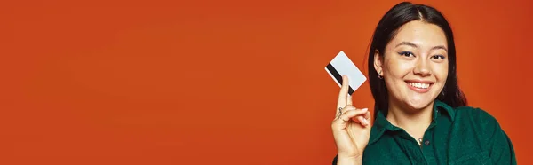 Heureux jeune asiatique femme en vert veste tenant carte de crédit sur fond orange, shopping bannière — Photo de stock