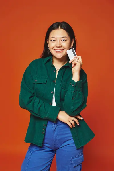 Positive jeune femme asiatique en veste verte tenant carte de crédit sur fond orange, consumérisme — Photo de stock