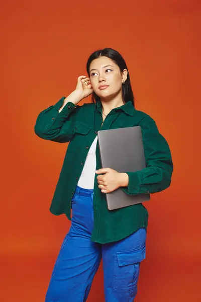 Pensivo jovem asiático mulher no vibrante traje segurando laptop no laranja fundo, remoto trabalho — Fotografia de Stock