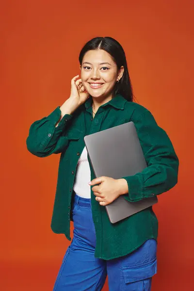Щаслива молода азіатка в яскравому вбранні тримає ноутбук на помаранчевому фоні, віддалена робота — стокове фото