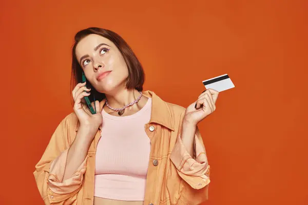 Pensive woman talking on smartphone and holding credit card on orange background, online shopping — Stock Photo