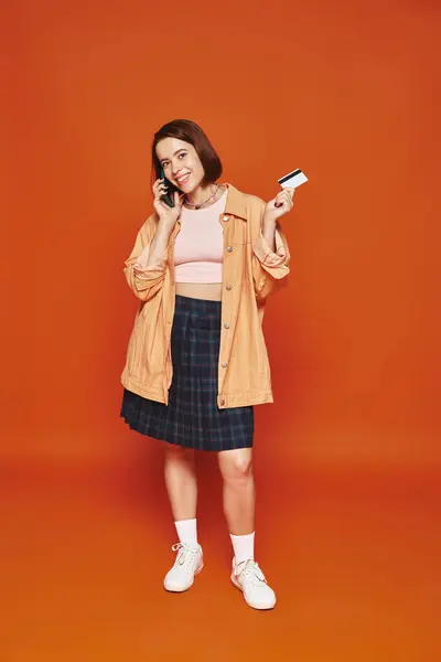 Щаслива жінка розмовляє на смартфоні і тримає кредитну картку на помаранчевому фоні, онлайн-покупки — стокове фото