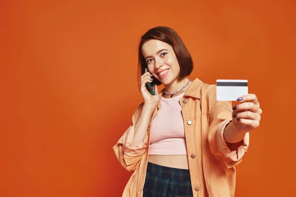 Happy woman talking on smartphone and holding credit card on orange background, online shopping — Stock Photo
