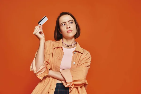 Pensive young woman with short hair holding credit card on orange background, personal finance — Stock Photo