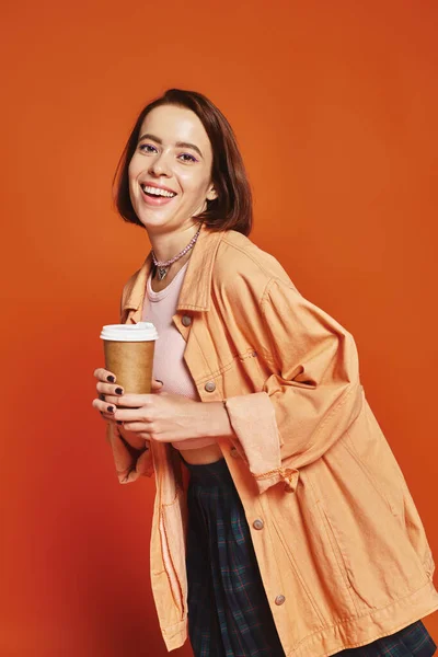 Longitud completa de la joven feliz con pelo corto sosteniendo taza de papel con café sobre fondo naranja - foto de stock