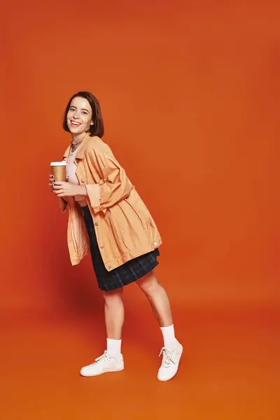 Alegre joven con pelo corto sosteniendo taza de papel con café mientras está de pie sobre fondo naranja — Stock Photo
