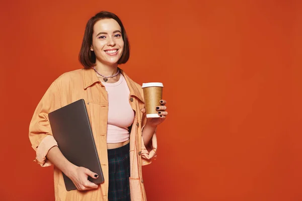 Веселая молодая женщина держит бумажную чашку с кофе и ноутбуком на оранжевом фоне, удаленная работа — стоковое фото