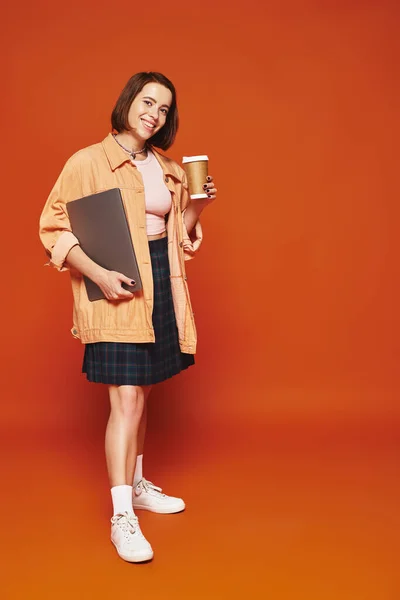 Joyeuse jeune femme tenant tasse en papier avec café et ordinateur portable sur fond orange, pigiste — Photo de stock