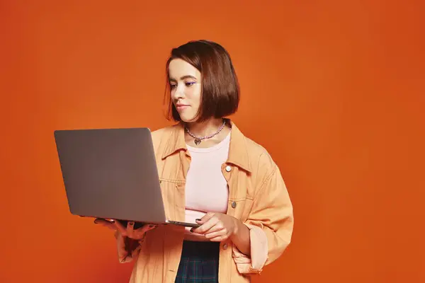 Молодой фрилансер в повседневной одежде держит ноутбук и работает удаленно на оранжевом фоне — стоковое фото