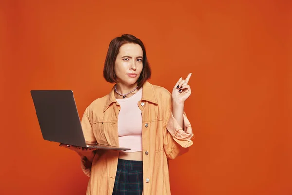 Giovane freelance in abbigliamento casual che tiene il computer portatile e punta con il dito su sfondo arancione — Foto stock