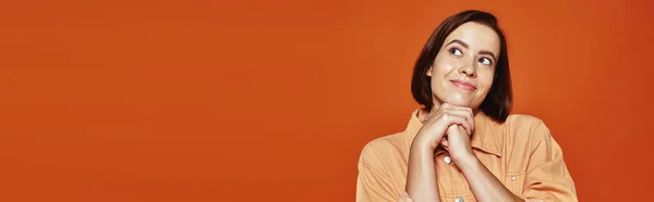 Надежная молодая женщина с короткими волосами улыбается и смотрит в сторону на оранжевом фоне, баннер — стоковое фото