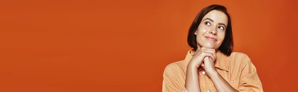 Mujer joven esperanzada con el pelo corto sonriendo y mirando hacia otro lado sobre fondo naranja, pancarta - foto de stock