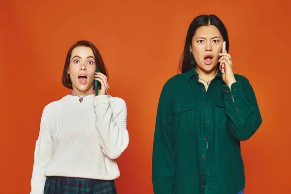 Dos impactadas mujeres multiculturales en traje vibrante hablando en teléfonos móviles sobre fondo naranja - foto de stock