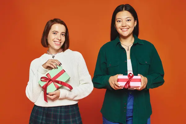 Happy young multicultural female friends holding presents on orange background, gift exchange — Stock Photo