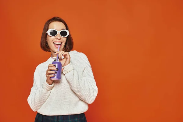 Stylish young woman in white trendy sunglasses sipping soda drink from can on orange backdrop — Stock Photo