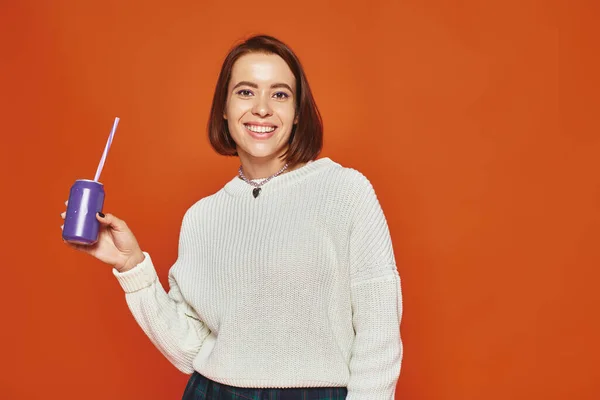 Cheerful young woman in white knitted sweater holding soda can with straw on orange backdrop — Stock Photo