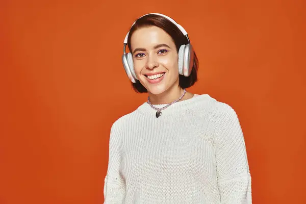 Fröhliche junge Frau in weißem Pullover und kabellosen Kopfhörern, die Musik auf orangefarbenem Hintergrund hört — Stockfoto