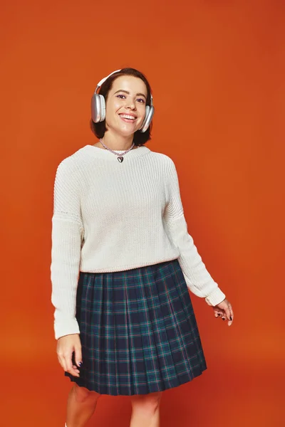 Joyful young woman in white sweater and wireless headphones listening music on orange backdrop — Stock Photo