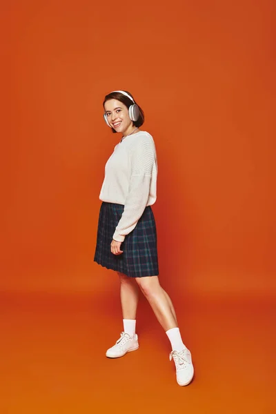 Jovem mulher positiva em suéter branco e fones de ouvido sem fio ouvindo música no fundo laranja — Fotografia de Stock