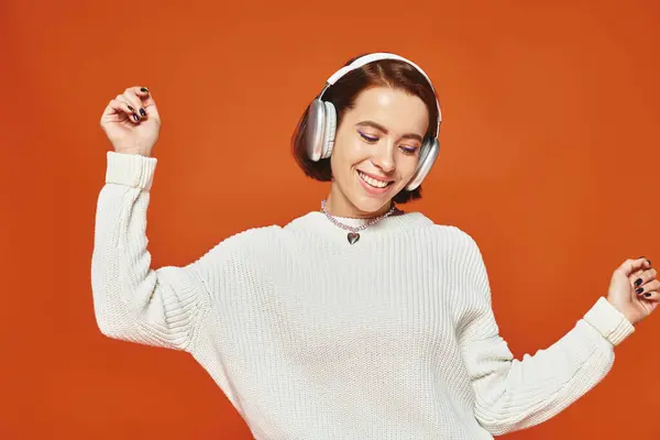Positive young woman in white sweater and wireless headphones enjoying music on orange backdrop — Stock Photo
