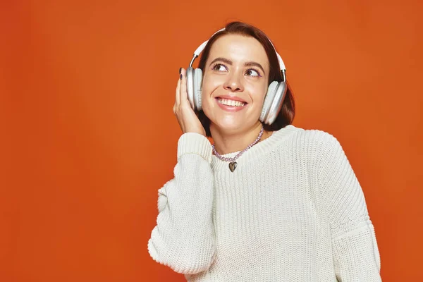 Mujer joven feliz en suéter blanco y auriculares inalámbricos disfrutando de la música en el fondo naranja - foto de stock