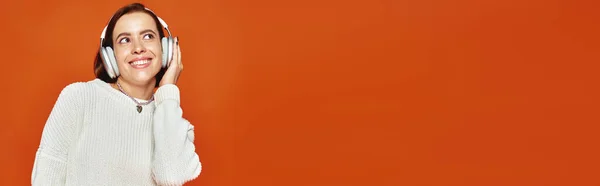 Весёлая женщина в белом свитере и беспроводных наушниках слушает музыку на оранжевом фоне, баннер — стоковое фото