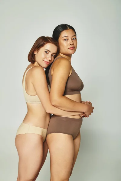 Dos mujeres multiétnicas en ropa interior abrazando y mirando a la cámara sobre fondo gris claro - foto de stock