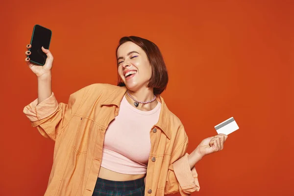 Mujer feliz joven sosteniendo teléfono inteligente y tarjeta de crédito sobre fondo naranja, concepto de comercio electrónico - foto de stock