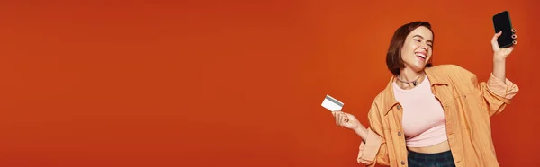 Mujer feliz joven sosteniendo teléfono inteligente y tarjeta de crédito sobre fondo naranja, banner de compras en línea - foto de stock