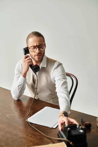 Good looking young man with glasses and beard sitting at table and talking by retro phone in office — Stock Photo