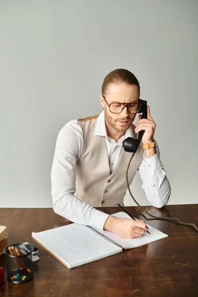 Handsome bearded man with glasses talking by retro phone and taking notes at table in office — Stock Photo