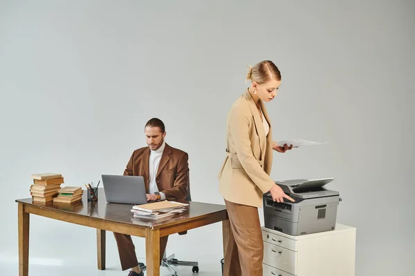 Young attractive woman in elegant suit using copy machine while her boyfriend working on laptop — Stock Photo