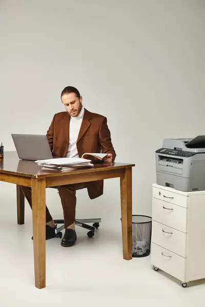 Привабливий бородатий чоловік в коричневій стильній куртці працює на ноутбуці і дивиться на паперові роботи — стокове фото