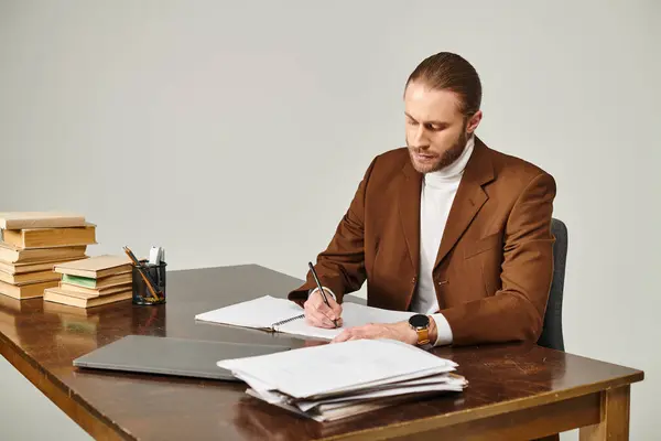 Joven guapo con barba en chaqueta marrón sentado en el escritorio y tomando notas con atención - foto de stock