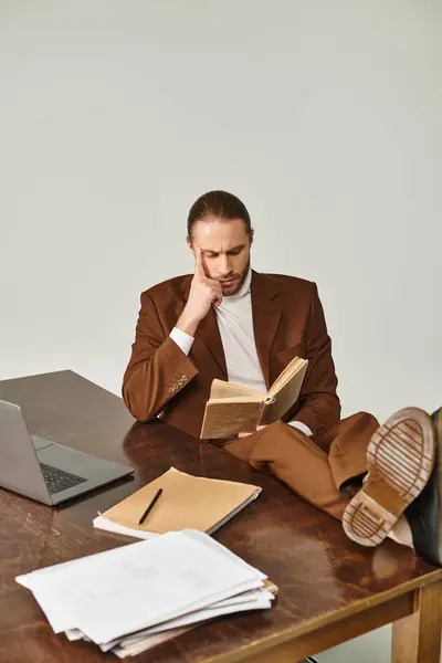 Concentrated young man with beard in elegant brown jacket reading book and posing with legs on table — Stock Photo