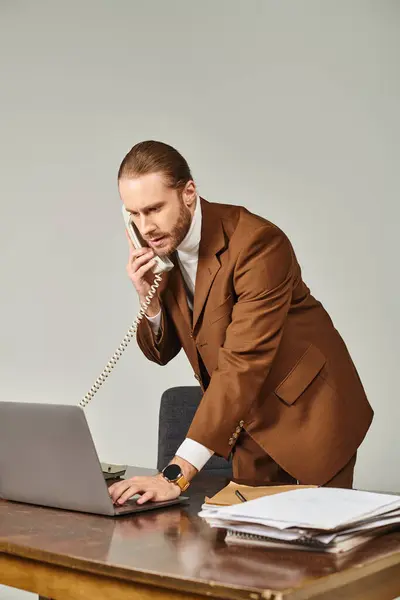 Hombre barbudo de buen aspecto en traje elegante hablando por teléfono retro y mirando a su computadora portátil en la oficina - foto de stock