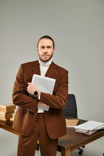 Beau jeune homme avec barbe dans une veste marron élégant tenant la paperasse et regardant la caméra — Photo de stock