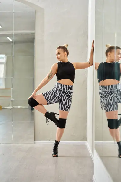 Mujer de moda en pantalones cortos de cebra y top balances negros en una pierna mientras baila contra el espejo - foto de stock