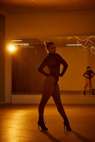 Una mujer equilibrada con mallas de rejilla y maillot negro se levanta contra un espejo de estudio en la pista de baile - foto de stock