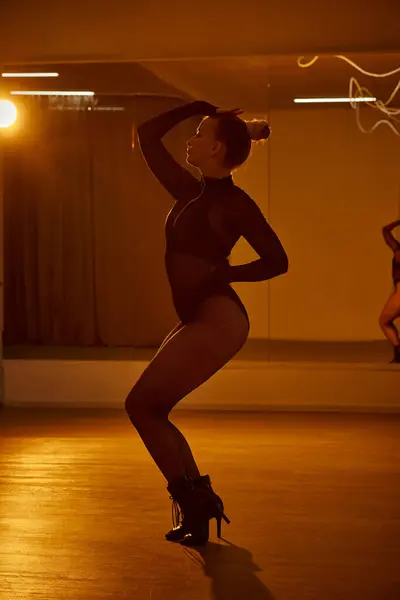 Bonita bailarina en medias de rejilla y maillot negro se levanta contra un espejo de estudio en la pista de baile - foto de stock