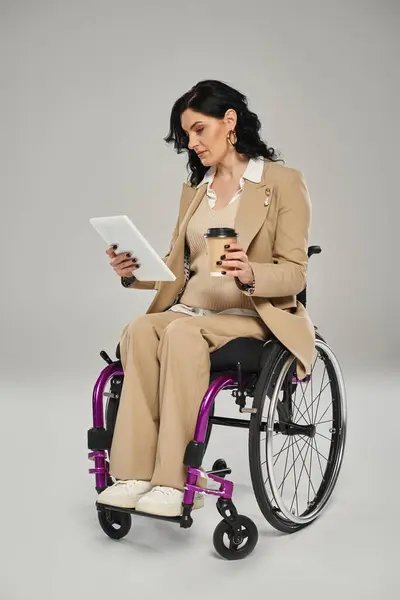 Good looking disabled woman in pastel chic attire in wheelchair looking at tablet and holding coffee — Stock Photo