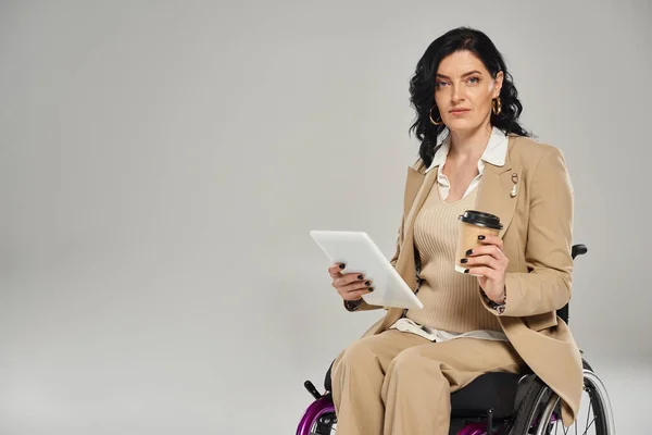 Atractiva mujer discapacitada en traje de pastel en silla de ruedas con café y tableta mirando a la cámara - foto de stock