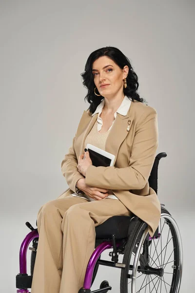 Mujer atractiva con discapacidad sentada en silla de ruedas y mirando a la cámara con la tableta en las manos - foto de stock