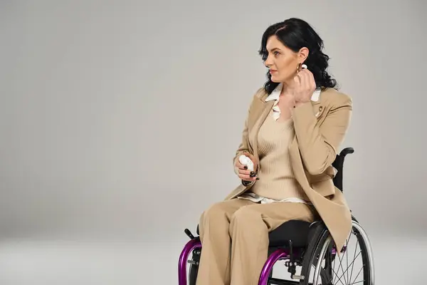 Hermosa mujer discapacitada en traje pastel en silla de ruedas poniéndose los auriculares sobre fondo gris - foto de stock