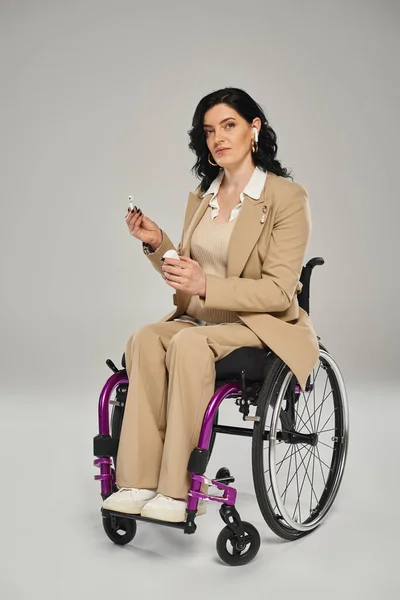 Mujer discapacitada con confianza en traje pastel en silla de ruedas poniéndose auriculares y mirando a la cámara - foto de stock