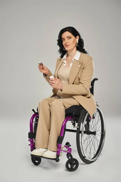 Confident disabled woman in pastel suit in wheelchair putting on headphones and looking at camera — Stock Photo