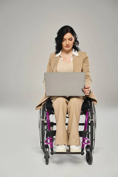 Atractiva mujer con discapacidad de movilidad sentada en su silla de ruedas y trabajando en su portátil - foto de stock