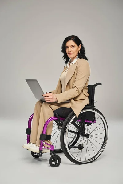 Mujer segura de buen aspecto con discapacidad en silla de ruedas con su computadora portátil mirando a la cámara - foto de stock